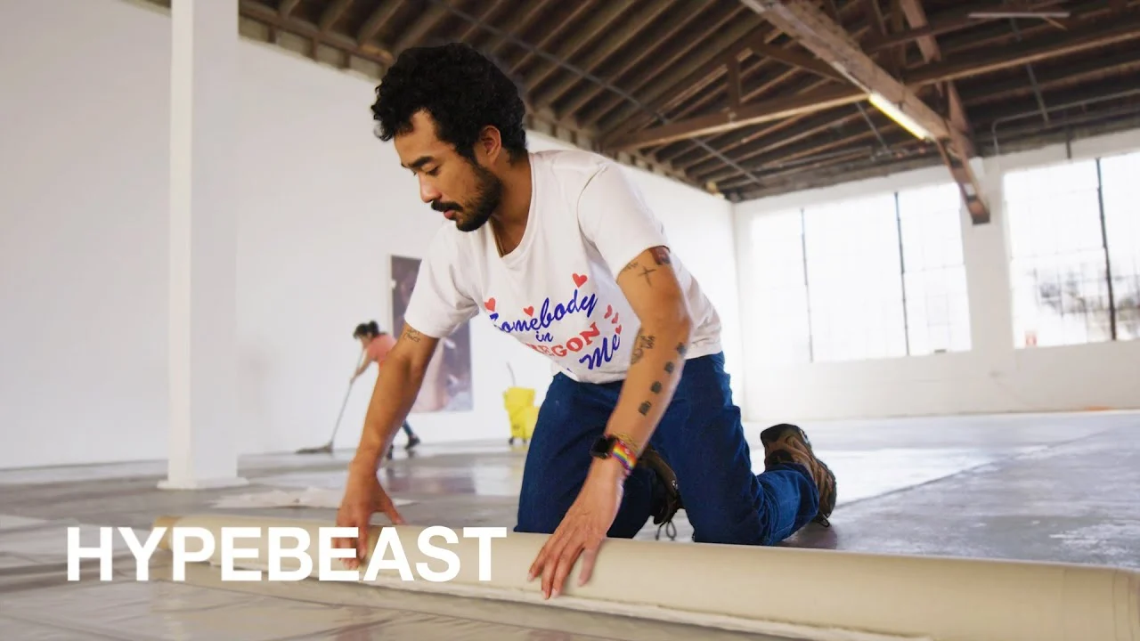 Lucien Smith kneeling to unroll canvas in an empty white studio, a frame from the HYPEBEAST Diaries interview carrying the publisher's watermark